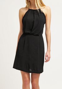 Robe noire à col halter en tissu léger, avec des plis au niveau du bodice, taille cintrée et jupe évasée qui tombe au-dessus des genoux.