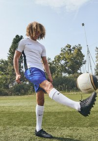 Nike hvid kortærmet skjorte med sorte detaljer, blå shorts, hvide sokker og sorte fodboldstøvler. Sparker en mønstret fodbold på græs.