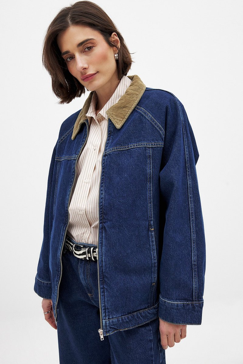 Tumši zila džinsa jaka ar korduroja apkakli. Iekļauj rāvējslēdzēju, kabatas ar plāksteriem un brīvu piegriezumu. Zem tās valkāta svītraina krekls.