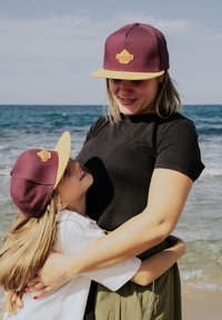 Dos personas con gorras similares: una color burdeos con visera amarilla que lleva un logo. Están en la playa, con el océano al fondo.