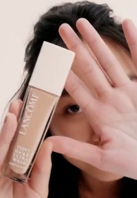 Lancôme-foundation i en glassflaske med hvit topp. Flasken viser en beige nyanse med preget tekst og en blank finish. Hånden skjuler delvis utsikten.