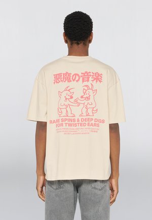 Person trägt ein beiges T-Shirt mit pinkfarbenen Cartoon-Teufeln und einem Text über seltene Musik-Singles auf der Rückseite, kombiniert mit grauen Jeans.