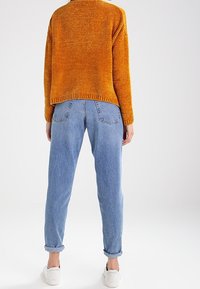 Oranger Strickpullover mit strukturierter Oberfläche, kombiniert mit hellblauen, hochgekrempelten Jeans und weißen Turnschuhen. Rückansicht gezeigt.