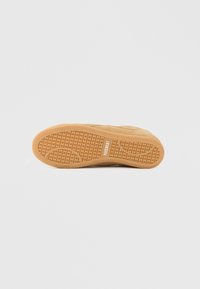 Semelle de sneaker en caoutchouc beige avec un motif de grip texturé et une tige en suède marron clair. Marquage notable en blanc au niveau de la voûte plantaire.