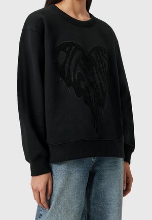 Personne portant un sweat-shirt noir avec un grand motif cœur texturé sur la poitrine et un jean bleu clair sur un fond uni.