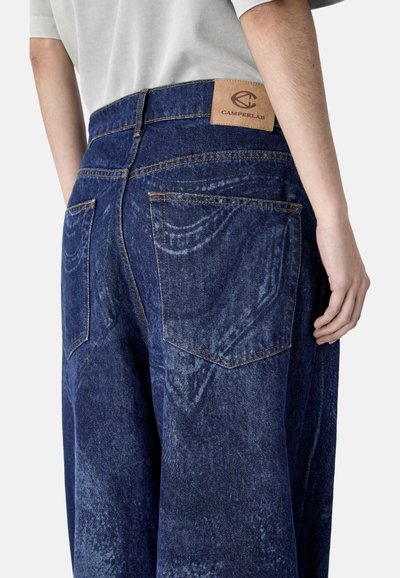 Person trägt locker sitzende dunkelblaue Jeans aus Denim mit detaillierter Naht und Camperlab-Logoaufnäher am Bund, darüber ist ein weißes Hemd sichtbar.
