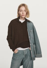 Persona bionda che indossa un maglione a trecce marrone scuro, una maglietta grigia chiara, jeans lunghi blu larghi e una giacca di denim oversize parzialmente drappeggiata.