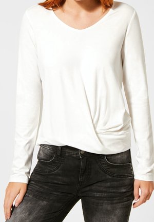 Wit top met lange mouwen en een geplooide zijkant, gecombineerd met vervaagde zwarte jeans, gedragen door een persoon met rood haar, bijgesneden ter hoogte van de nek en heupen.