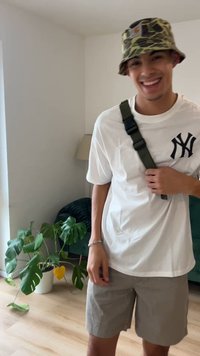 Balta īsroku T-krekla ar melnu NY logotipu, pelēki šorti un kamuflāžas cepure ar plata malu. Tur zaļas siksnas somu. Fonā ir redzami augi.