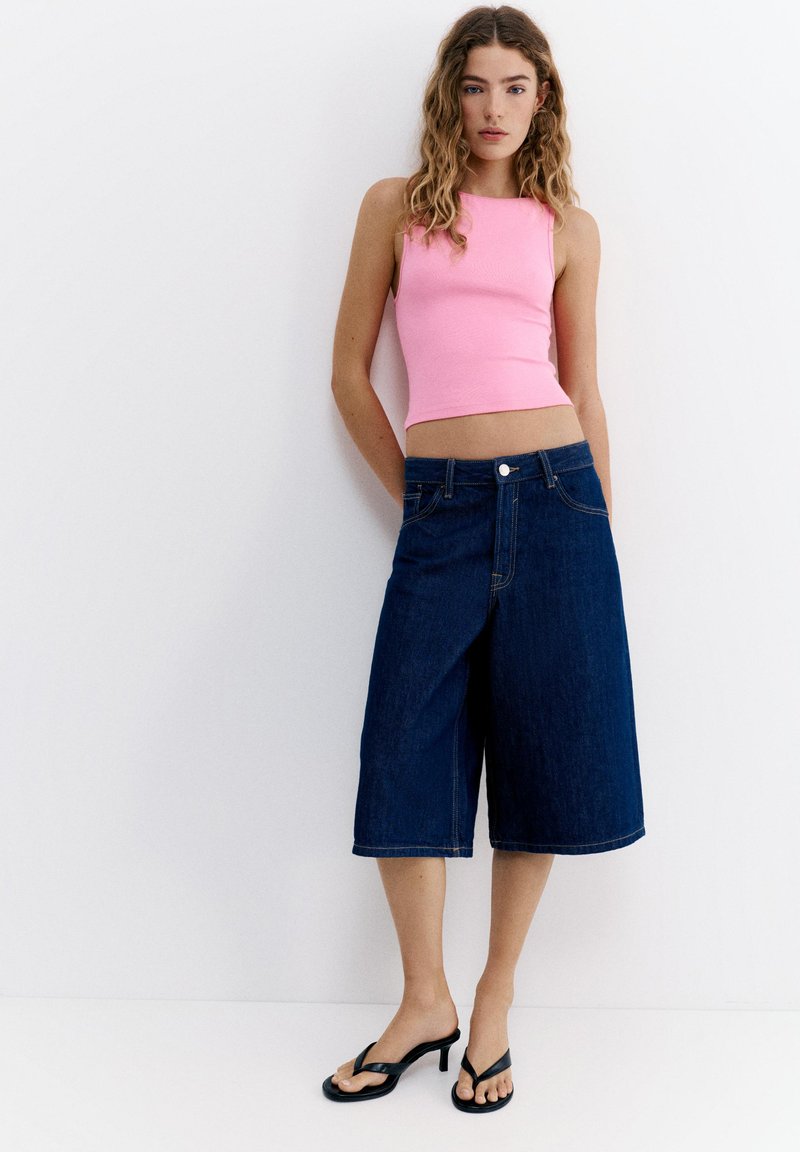Jeune femme portant un crop top rose sans manches, un pantalon large en denim bleu foncé, et des sandales noires à talons, debout contre un mur blanc.