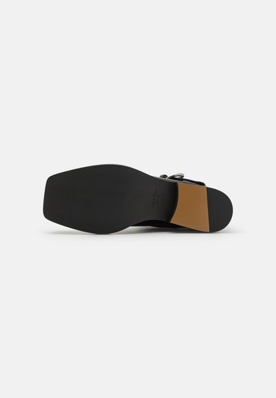 Černá obuv s širokou plochou podrážkou a hnědým zdobením na patě. Má hladký povrch a minimalistický design.
