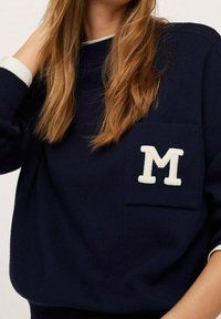 Jersey de punto azul marino con un parche blanco en forma de "M" en el bolsillo del pecho y un cuello interior blanco en contraste. Tejido de textura suave.