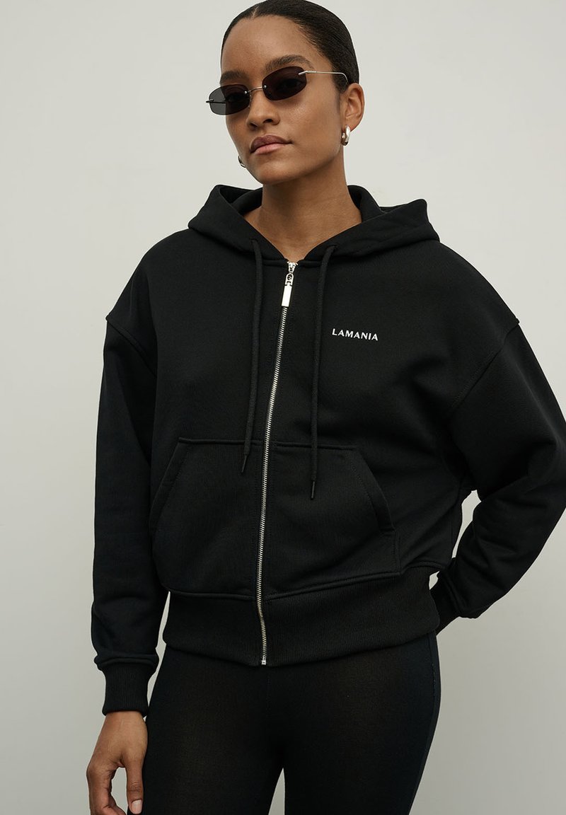 Juodas su gobtuvu džemperis su priekine užtrauktuko uždarymo sistema, didelė kengūros kišenė ir balta „LAMANIA“ logotipas. Tekstūruota medžiaga, laisvas siluetas ir grublėti rankogaliai.