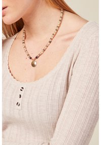 Collana di perline con perline chiare e scure e un pendente circolare tono oro. Chi la indossa ha una maglietta rigata di colore chiaro con bordi ondulati.