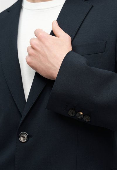 Χέρι που προσαρμόζει το πέτο ενός σκούρου μπλε σακακιού κουστουμιού πάνω από ένα λευκό μπλουζάκι με στρογγυλή λαιμόκοψη, εστιάζοντας σε κουμπιά και υφή υφάσματος.