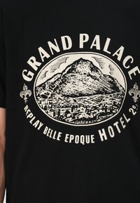 T-shirt nera con una grafica illustrata bianca di una montagna con uno skyline, circondata da testo circolare 'GRAND PALACE' e 'REPLAY BELLE EPOQUE HOTEL.'