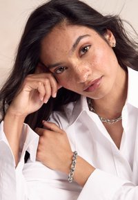 Silver chain necklace and bracelet worn over a white shirt, showcasing smooth metal links and a close-up of a hand resting on the face.