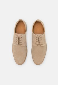 Chaussures beige en daim à lacets avec un bout arrondi, semelle plate et détails cousus. Elles présentent une semelle intérieure en cuir lisse avec un branding en relief.