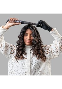 Krølltang med svart ståltopp, holdt over hodet. Modellen har på seg en hvit, åpen bluse. Svart beskyttelseshanske på den ene hånden. Grå bakgrunn.