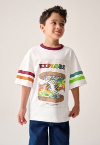 Fehér, túlméretezett póló bordó, narancssárga és zöld csíkokkal az ujjain, valamint egy színes cipőillusztrációval, amely terepmintákat ábrázol.