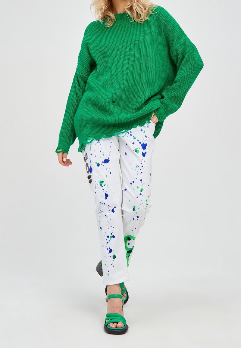 Personne portant un pull en maille vert extra-large aux bords usés, un pantalon blanc avec des éclaboussures de peinture bleue et verte, et des sandales vertes à talons.