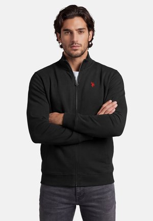 Mann mit verschränkten Armen, der eine schwarze Zip-Jacke mit rotem, aufgesticktem Logo auf der Brust und graue Jeans trägt.