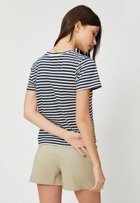 Donna con capelli lisci e scuri, che indossa una maglietta corta a righe blu navy e bianche e pantaloni corti beige, in piedi su uno sfondo neutro.