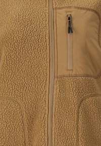 Vaaleanruskea fleece-takki, jossa on teksturoitu kangas, keskellä vetoketju ja sileä vetoketjullinen rintatasku mustilla vetonuppiloilla.