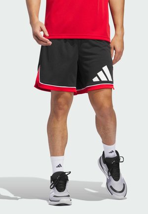 Person trägt schwarze und rote Sportshorts mit weißem Logo, weiße Socken und graue sowie schwarze Sportschuhe und steht auf hellem Hintergrund.