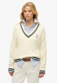 Cremfarbener V-Ausschnitt-Pullover mit grünen Streifenakzenten. Rippenbündchen und Saum. Über einem blau-weißen gestreiften Hemd mit Knöpfen getragen. Leichter Stoff.