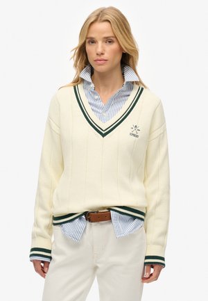 Suéter crema de cuello en V con acentos de franjas verdes. Puños y dobladillo acanalados. Usado sobre una camisa de botones a rayas azul y blanca. Tela ligera.