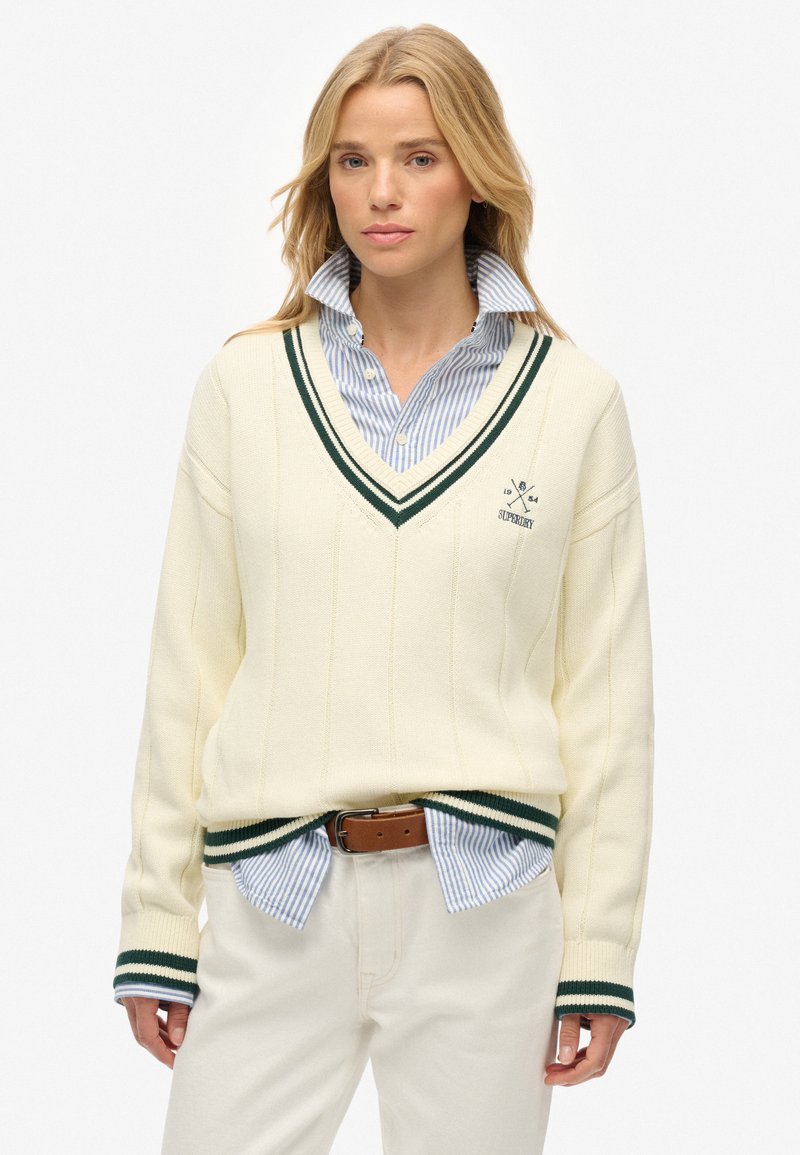Cremfarbener V-Ausschnitt-Pullover mit grünen Streifenakzenten. Rippenbündchen und Saum. Über einem blau-weißen gestreiften Hemd mit Knöpfen getragen. Leichter Stoff.