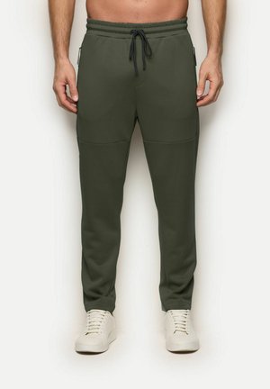 Mężczyzna ubrany w oliwkowozielone spodnie dresowe ze ściągaczem i białe sneakersy, stojący na jednolitym tle.