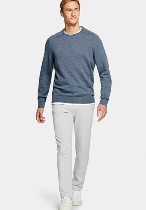 Man loopt, draagt een blauwe trui met lange mouwen, wit hemd, lichtgrijze broek en witte sneakers op een effen lichte achtergrond.