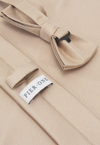 Beiger gewebter Stoff mit einem kleinen schwarzen Verschluss und einem weißen Etikett mit der Aufschrift "PIER · ONE." Der Stoff hat ein gewebtes Muster und eine glatte Oberfläche.