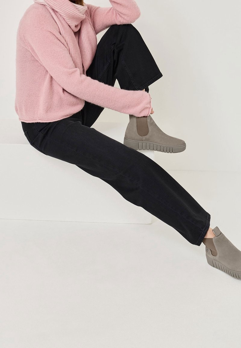 Personne portant un pull rose en maille pelucheuse et un pantalon noir, assise avec un genou relevé, montrant des bottines grises à semelles épaisses.
