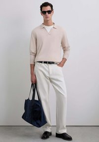 Un pull en tricot beige clair avec un col, un pantalon en denim blanc, une ceinture noire et des chaussures noires, tenant un sac fourre-tout bleu marine.