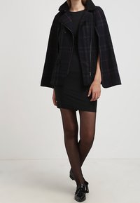 Veste style cape à carreaux noirs avec fermeture éclair à l'avant et col doux, superposée sur une robe noire ajustée et des collants noirs transparents. Chaussures plates.