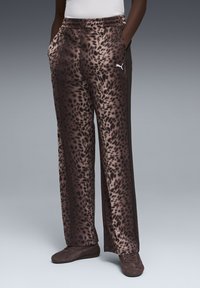 Άτομο που φοράει καφέ φόρμα Puma με σχέδιο λεοπάρδαλης και ταιριαστά καφέ αθλητικά παπούτσια Puma, στέκεται με τα χέρια στις τσέπες.