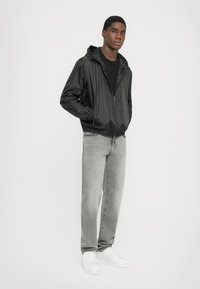 Svart huva jacka med kviltat mönster, kombinerad med grå jeans och vita sneakers. Tyget verkar lätt och glansigt, med elastiska ärmslut.