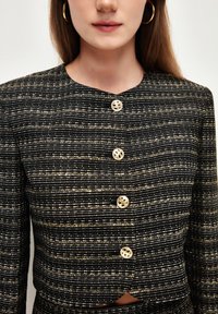 Blazer a rayas negro y dorado con escote redondeado, que presenta botones metálicos y un diseño ajustado. Tejido texturizado con un ligero brillo.