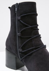 Bottine en suede noire avec un talon épais, featuring des lacets décoratifs et une fermeture éclair sur le côté pour un enfilage facile. La finition texturée ajoute de la profondeur.