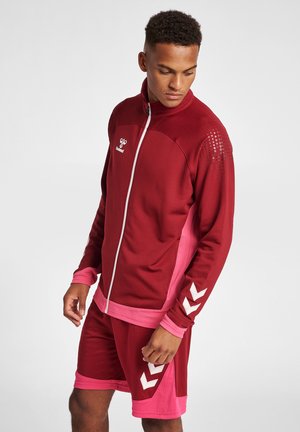 Młody mężczyzna patrzy w dół, nosząc czerwoną i różową sportową kurtkę na zamek oraz dopasowane spodenki z białymi akcentami w kształcie jodełki.