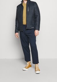 Svart, vadderad jacka med hög krage över en senapsgul T-shirt, kombinerad med marinblå byxor och bruna högstövlar. Dragkedjefickor som detalj.