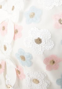 Λευκό ύφασμα με πολύχρωμα κεντημένα λουλούδια σε ροζ, μπλε και λευκό, το καθένα με κεντρική χρυσή λεπτομέρεια. Διαθέτει λεπτομέρεια με φιόγκο.