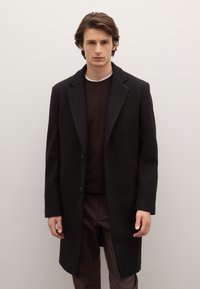 Abrigo negro con solapa de muesca, cierre de un solo botón y bolsillos laterales, combinado con un suéter marrón, sobre un fondo claro y sencillo.