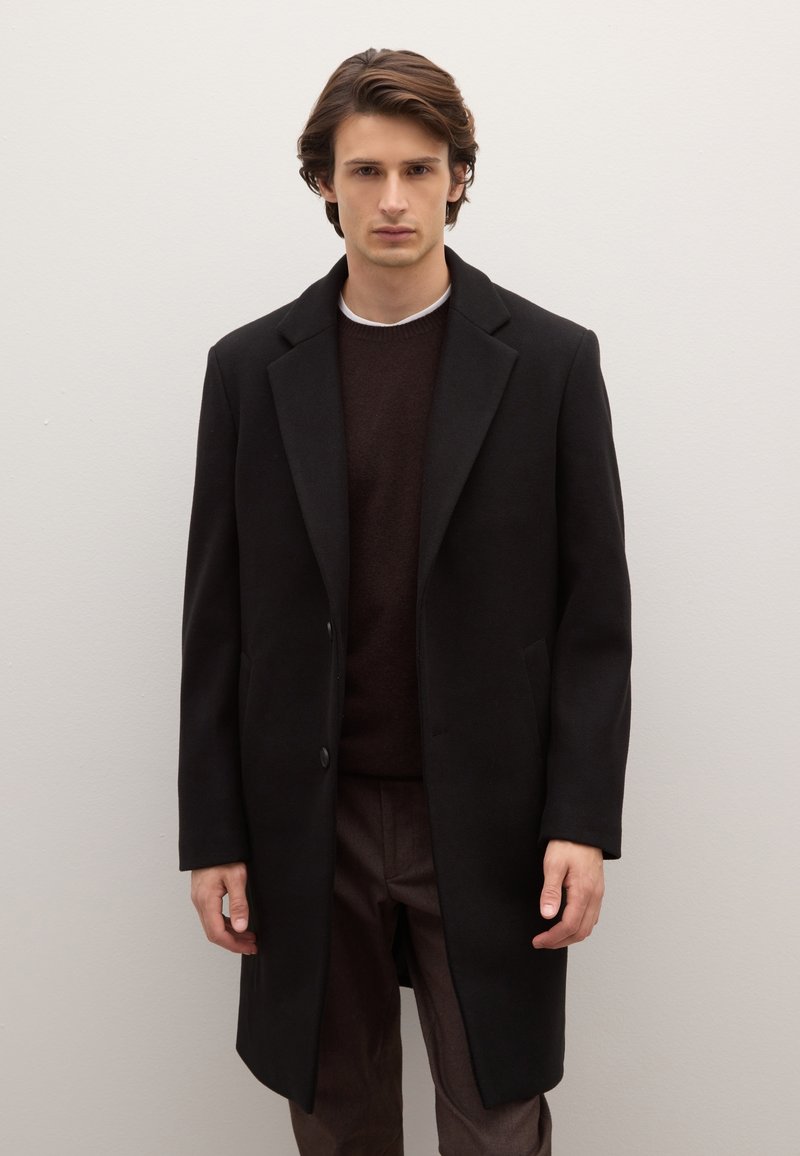 Abrigo negro con solapa de muesca, cierre de un solo botón y bolsillos laterales, combinado con un suéter marrón, sobre un fondo claro y sencillo.