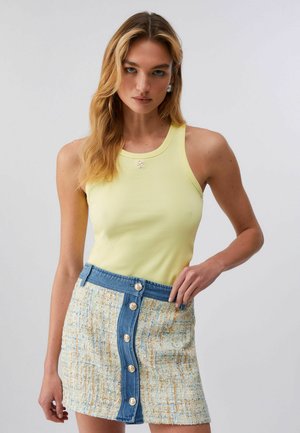 Donna con lunghi capelli biondi indossa un top giallo senza maniche a costine e una minigonna texturizzata con patta di bottoni in denim blu, in piedi davanti a uno sfondo semplice.