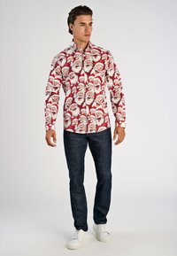Rode button-up shirt met een herhaald patroon van een kerstman gezicht. De shirt heeft lange mouwen en wordt gecombineerd met donkere spijkerbroek en witte sneakers.