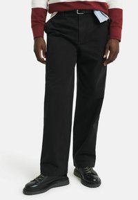 Schwarze Hose mit entspannter Passform, aus Baumwolle gefertigt. Mit schräge Taschen und Gürtelschlaufen. Kombination mit schwarzen Schnürschuhen.